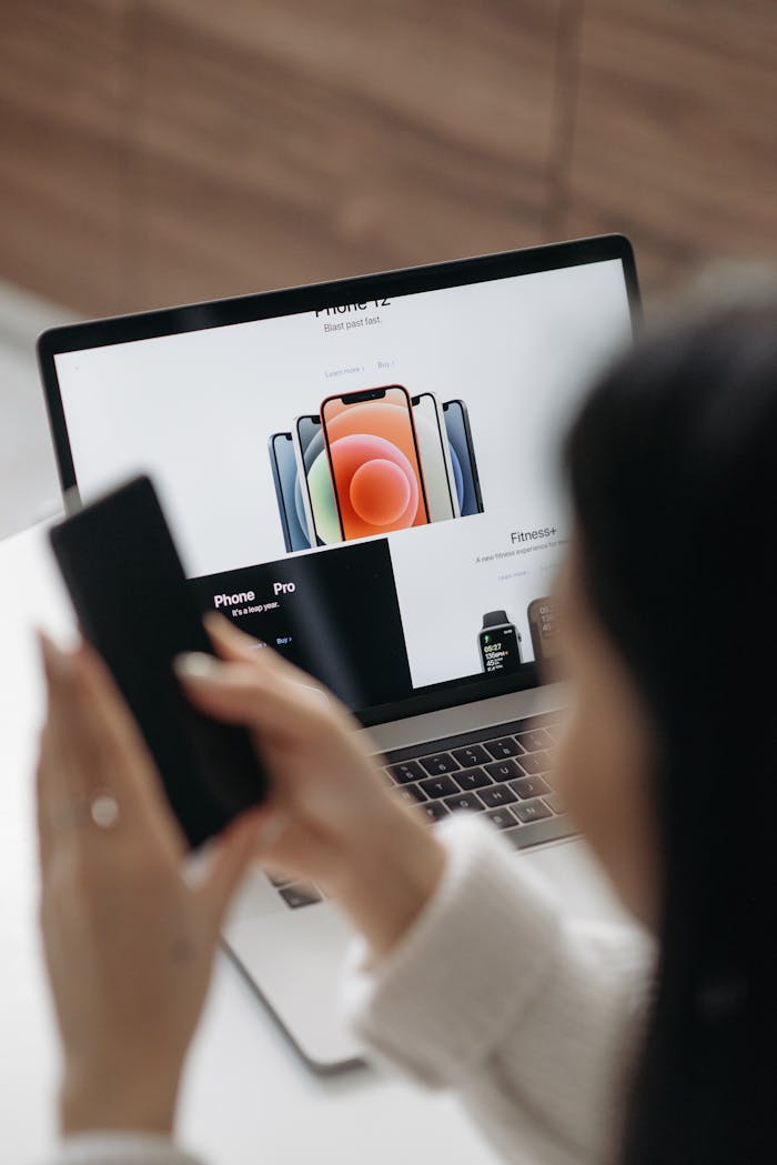 Woman using a laptop and smartphone to browse electronics indoors.